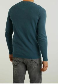 Man wearing teal long-sleeve textured shirt and faded gray jeans, standing with back facing the camera against a plain background.