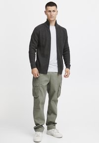 Dunkelgrauer, gerippter Reißverschluss-Pullover, darunter ein weißes Rundhals-Shirt, olivgrüne Cargo-Hosen mit großen Taschen und weiße Sneakers.