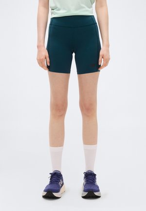 Persoon die een groene sportbroek, witte sportsokken en blauwe hardloopschoenen draagt, staand tegen een effen achtergrond.