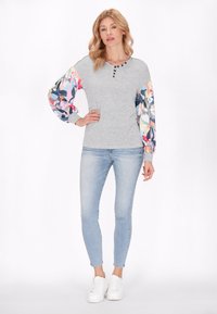 Szürke, hosszú ujjú felső színes virágmintás puff ujjakkal, gombos részletekkel a nyaknál, világoskék skinny farmerral és fehér túrázócipővel párosítva.