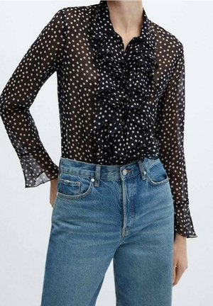 Blouse noire transparente à manches longues avec pois blancs et volant sur le devant, associée à un jean denim bleu taille haute.