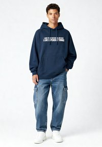 Sweat à capuche bleu marine avec texte blanc, poche kangourou et capuche à cordon, associé à un jean cargo bleu clair et des baskets blanches.