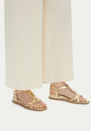 Piedi con sandali piatti in metallo dorato con cinturini alla caviglia e dettagli con borchie, abbinati a pantaloni larghi color crema.