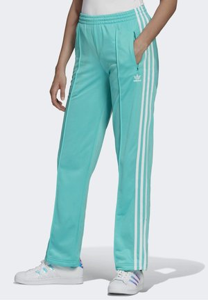 Person iført turkise Adidas træningsbukser med hvide striber og matchende Adidas sneakers, står med den ene hånd i lommen.