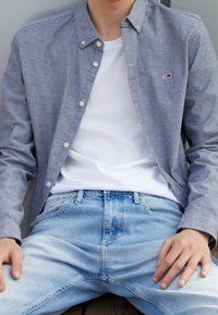 Button-up shirt in light gray fabric, worn over a white t-shirt. Light blue jeans with a faded texture. Hands positioned on thighs.
