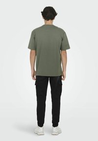 Olive green cotton t-shirt with short sleeves, paired with black cargo pants featuring side pockets and elastic cuffs, worn with white sneakers.