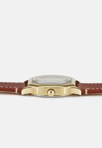 Montre dorée avec un boîtier carré, bracelet en cuir marron, couture crème et couronne ronde. Cadran en verre avec un motif concentrique visible.