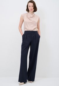Beige, ärmlös blus med draperad halsringning, som kombineras med högmidjade, marinhäftiga vida byxor. Slät textur med skräddarsydda veck.