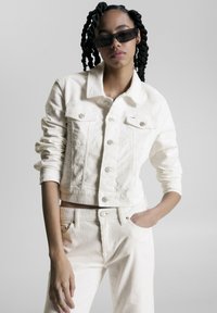 Chaqueta corta de pana blanca con cierre de botones, dos bolsillos frontales y pantalones de pana blanca a juego. La modelo lleva gafas de sol oscuras.