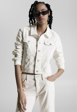Veste cropped en velours côtelé blanc avec fermeture à boutons, deux poches avant et pantalon en velours côtelé blanc assorti. Le mannequin porte des lunettes de soleil noires.