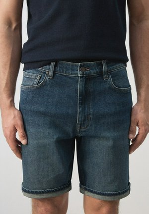 Hombre con pantalones cortos vaqueros azul oscuro con dobladillos enrollados y camisa azul marino, de pie con los brazos relajados a los lados.