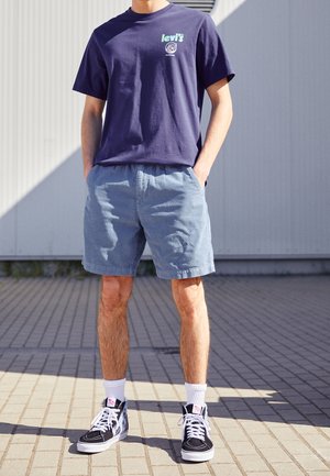 Mann trägt ein dunkelblaues Levi's T-Shirt, hellblaue Shorts, weiße Socken und schwarze High-Top-Sneakers und steht im Freien auf gepflastertem Boden.