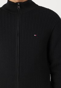 Svart zip-up tröja med hög krage, med ett texturerat fiskbensmönster. Inkluderar en liten logotyp i rött, vitt och blått på bröstet.