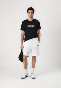 Svart T-skjorte med hvit "JACK & JONES" logo, grå shorts, svart bag, grå og hvite sneakers, og hvite ankelsokker. Uformell stil.