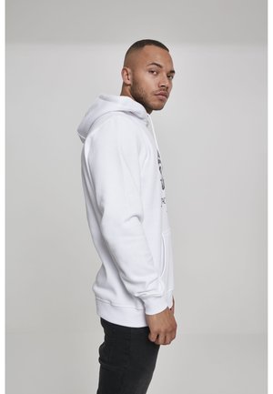 Uomo con capelli corti che indossa una felpa bianca con cappuccio e jeans neri, in piedi di profilo contro uno sfondo grigio chiaro uniforme.