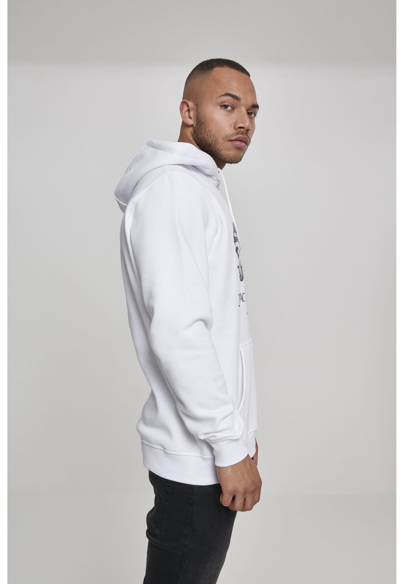 Homme aux cheveux courts portant un sweat à capuche blanc et un jean noir, debout de profil sur un fond gris clair uni.