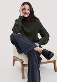 Donkergroene turtleneck trui, raglanmouwen, gecombineerd met marineblauwe corduroypantalons en zwarte franjesloafers, zittend op een textuurrijke crèmewitte stoel.