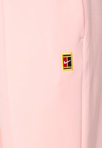 Ljust rosa tygjoggers med en broderad svart, röd och gul logotyp på vänster sida, med en slät yta och rena sömmar.