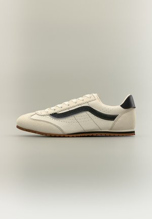 Άσπρο sneaker χαμηλού προφίλ με μαύρη πλευρική ρίγα, ανώτερο μέρος από μπεζ σουέτ, καφέ καουτσούκ σόλα και μαύρο ταμπ επιφάνειας φτέρνας, εμφανίζεται σε πλάγια προβολή.