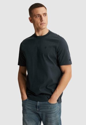 Homme aux cheveux courts et bruns, portant un t-shirt bleu marine foncé et un jean bleu, les mains dans les poches, regardant légèrement vers la droite.