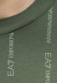 Κοντινό πλάνο πράσινου πλεκτού υφάσματος με ανοιχτόχρωμο κείμενο που γράφει «EMPORIO ARMANI EA7» κοντά σε ριμπ ύφασμα για γιακά.