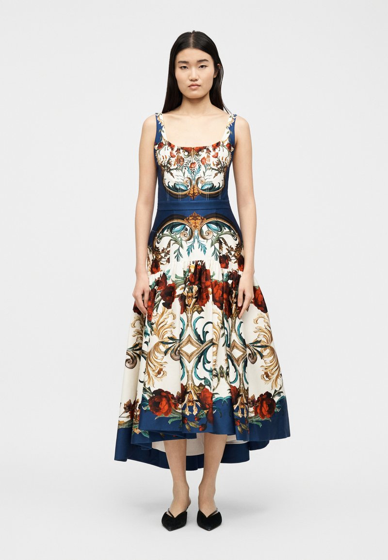 Femme portant une robe sans manches, asymétrique, avec des motifs floraux bleus, crème, rouges et des motifs dorés ornés, se tenant devant un fond blanc uni.