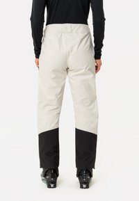 Person, die zweifarbige Ski-Hosen in Beige und Schwarz sowie schwarze Skistiefel trägt und mit dem Rücken zur Kamera vor einem weißen Hintergrund steht.