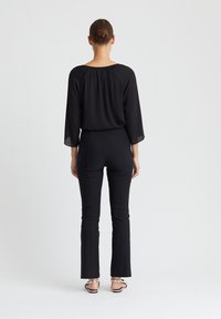 Frau mit hochgestecktem Haar, die ein lockeres schwarzes Oberteil, enge schwarze Hosen und silberne Sandalen trägt und vor einem schlichten weißen Hintergrund steht.