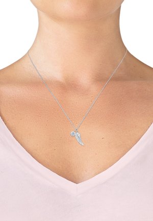 Silberne Halskette mit einem Flügel-Anhänger und einem kleinen klaren Edelstein-Anhänger, der über einem V-Ausschnitt-Oberteil mit glatter Textur ruht.