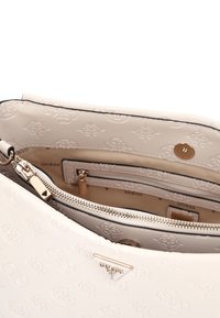 Bolso Guess blanco con relieve, cremallera dorada y logo, mostrando bolsillos interiores abiertos y cierre magnético.