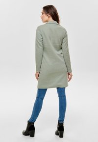 Cappotto lungo verde chiaro con maniche lunghe e colletto, texture morbida e vestibilità rilassata, abbinato a jeans blu e stivaletti neri con tacco.