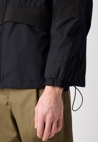 Den sorte jakke har en glat tekstur med elastiske manchetter, en lynlåslukning foran og et kontrasterende paneldesign. Stylet med khaki bukser.