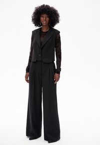 Czarny bezrękawnik na jeden guzik, w połączeniu z szerokimi spodniami. Bluzka ma przezroczyste długie rękawy z kwiatowym haftem.