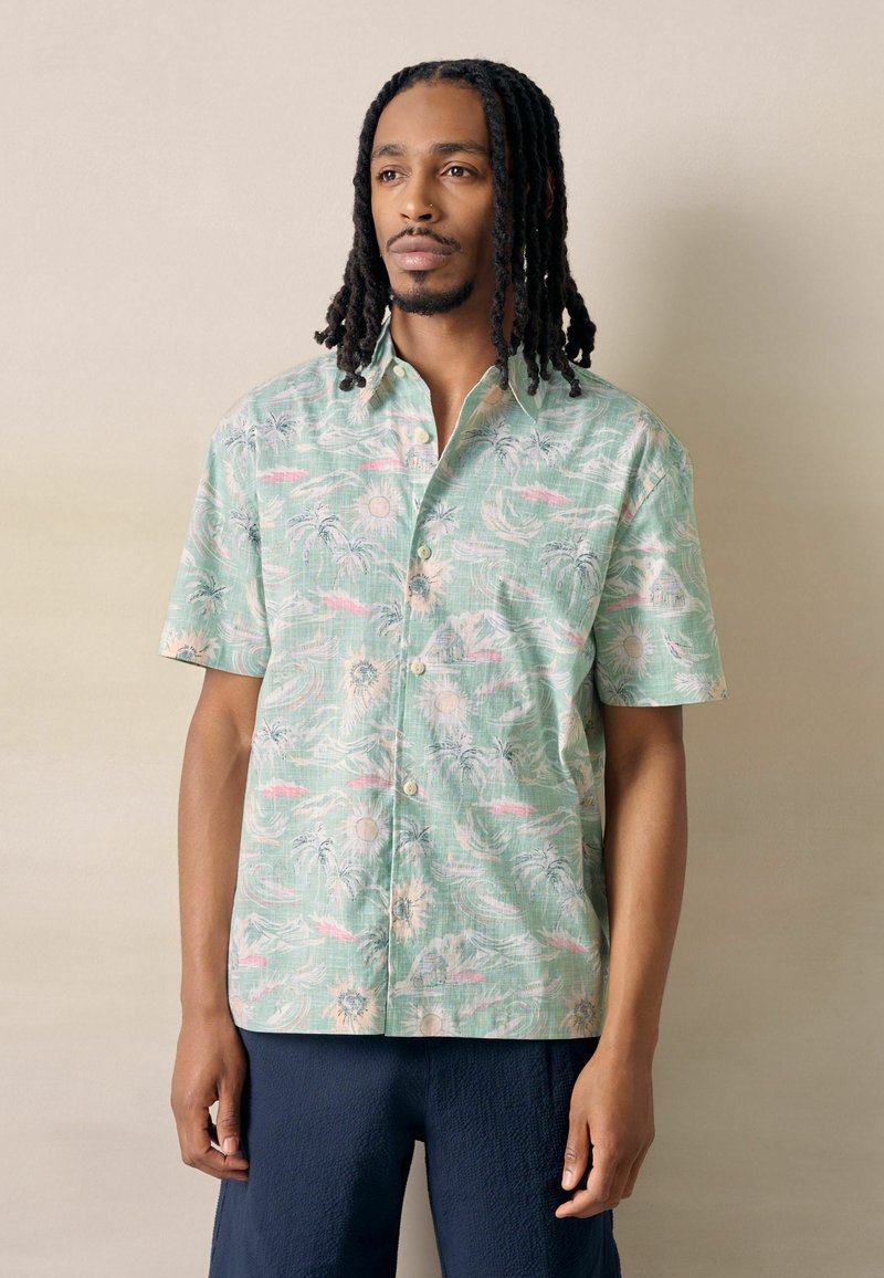 Homme aux dreadlocks mi-longs portant une chemise à manches courtes vert clair à boutons avec des motifs de palmiers et de soleil, debout devant un fond beige.