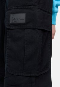 Black cargo pants made of durable fabric, featuring a prominent pocket, textured surface, and a sewn black logo patch.