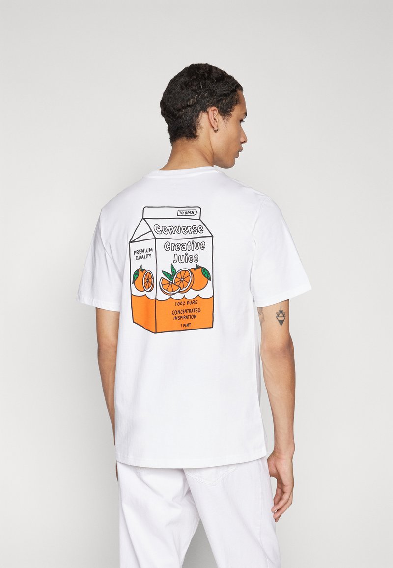 Camiseta blanca de algodón con un estampado gráfico de un cartón de zumo de naranja en la parte trasera. El texto incluye "Converse" y "Creative Juice."