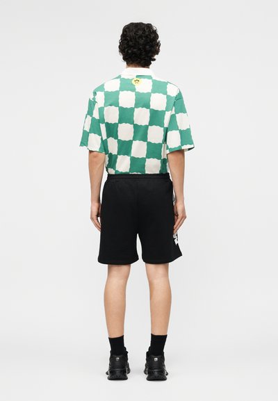 Muž otočený chrbtom, oblečený v zelenej a bielo kockovanej košeli s krátkym rukávom, čiernych šortkách, čiernych ponožkách a čiernych teniskách na jednoliatom pozadí.