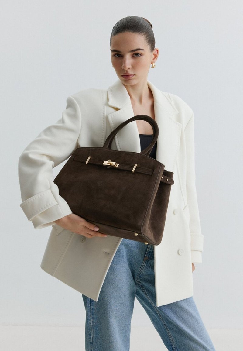 Donna con capelli tirati indietro indossa una giacca bianca e jeans, tenendo una grande borsa in suede marrone scuro con chiusura dorata su sfondo semplice.
