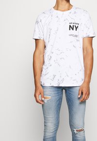 T-shirt i vitt marmormönster med rund halsringning, svart text "UP STATE NY", ihop med ljusblå slitna smala jeans.