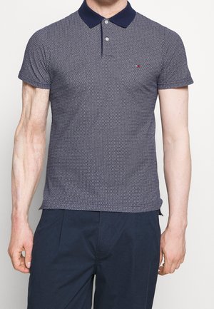 Mand iført en kortærmet navy mønstret poloshirt med en mørk krave og mørkeblå bukser, stående mod en ensfarvet baggrund.