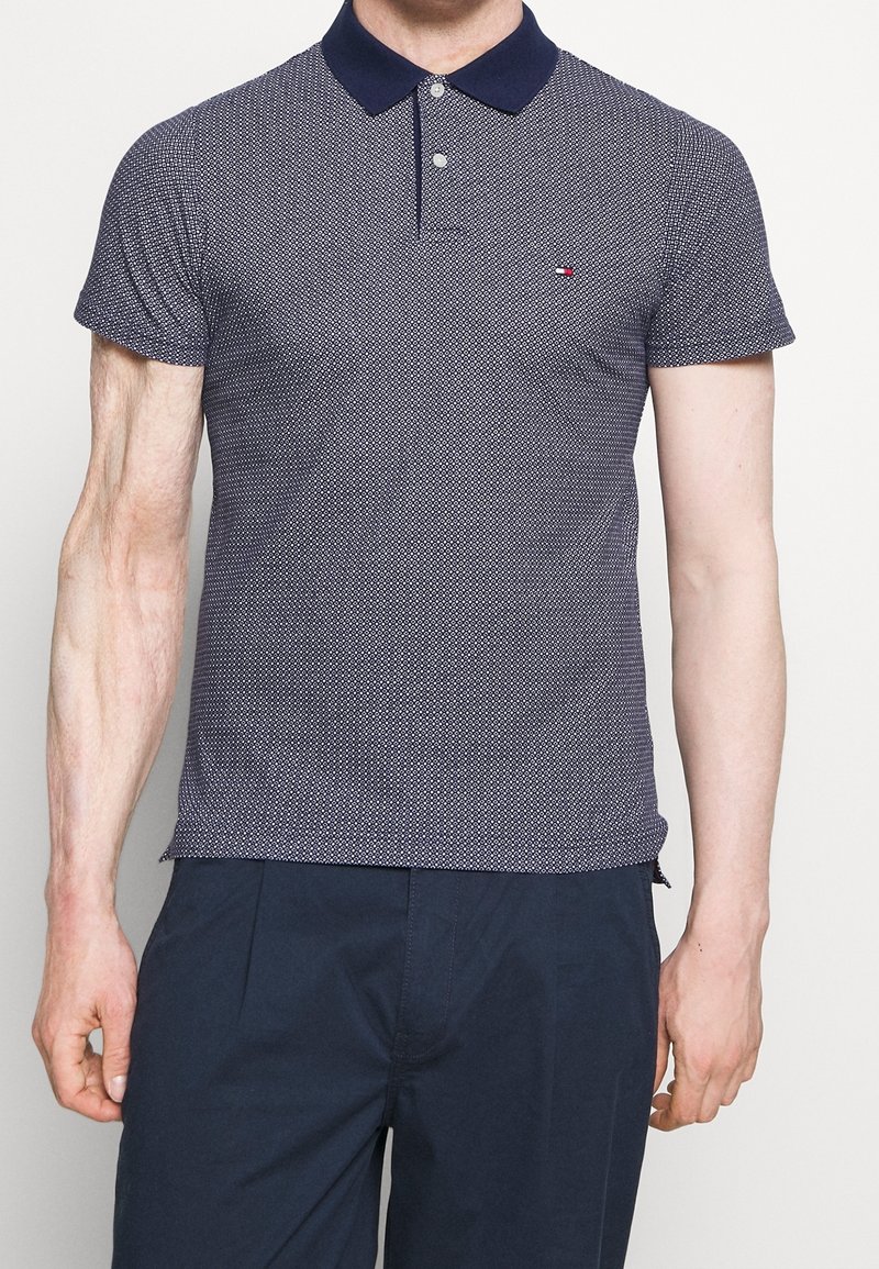 Homme portant un polo à manches courtes bleu marine à motifs avec un col sombre et un pantalon bleu foncé, debout devant un fond uni.