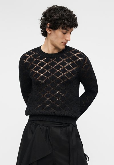 Νεαρός άνδρας με σγουρά μαλλιά που φοράει μαύρο διάφανο πουλόβερ με σχέδιο ρόμβους και φαρδιά μαύρη παντελόνια, κοιτάζει προς τα κάτω προς την αριστερή του πλευρά.