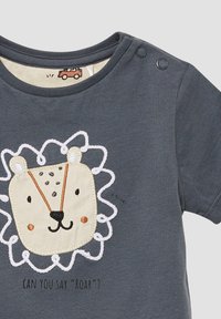 Szary t-shirt z krótkim rękawem z nadrukiem w postaci lwa, haftowanymi akcentami i napami na ramieniu. Tekst brzmi: "Czy potrafisz powiedzieć 'ryczeć'?"