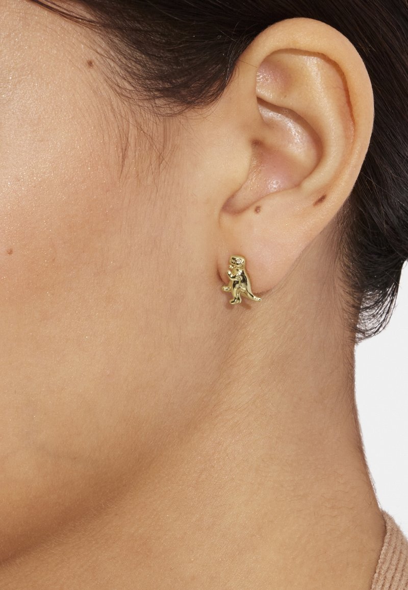 Petite boucle d’oreille clou en forme de dinosaure dorée, portée sur le lobe gauche de l’oreille sur une peau claire et des cheveux foncés.