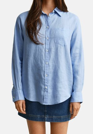 Femme portant une chemise bleu clair boutonnée avec une poche poitrine et une jupe en jean bleu foncé, debout devant un fond uni.