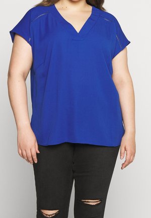Woman wearing a royal blue V-neck short-sleeve blouse with subtle stitch details paired with black ripped jeans.