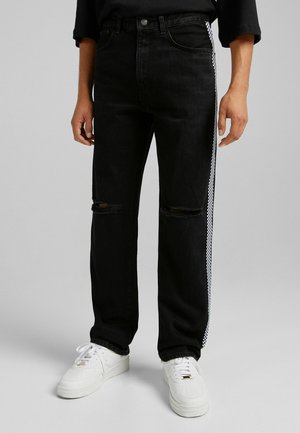 Persona vistiendo jeans negros desgastados con rayas laterales en blanco y negro a cuadros y zapatillas blancas sobre un fondo gris.