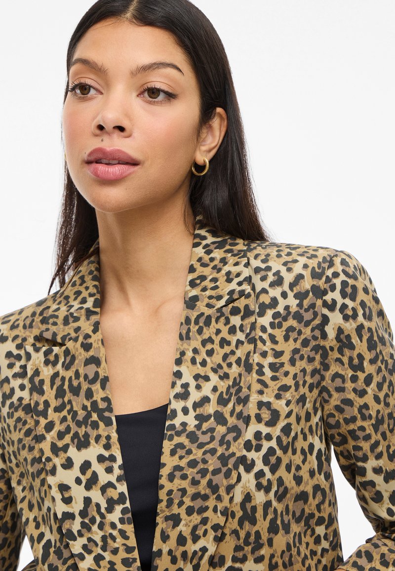 Leopardenmuster-Blazer in Beige und Schwarz, mit geschlitztem Kragen und glatter Textur. Trägt ein schwarzes Oberteil mit großen Ohrringen.