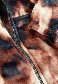 Bruine en zwart gemêleerde fleecejas met een zwarte rits en een stoffen textuur. Close-up toont zacht, pluche materiaal en een duidelijke verkleuring.