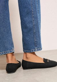 Černé semišové baleríny s plochou podrážkou a zlatým detailem vpředu, v kombinaci s modrými krátkými džínami s roztřepenými okraji.